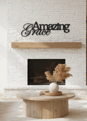 A modern living room with a white brick fireplace featuring black metal wall art that reads 'Amazing Grace'. The room is styled with two light beige sofas, a round wooden coffee table, and built-in wooden shelves on the right.