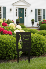 A black metal garden stake sign with a wine glass silhouette and the words 'Gnome Sweet Gnome 1308' displayed in a landscaped garden in front of a white house.