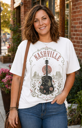 A woman wearing a white graphic t-shirt with a vintage-style 'Nashville' guitar design, paired with blue jeans and a brown shoulder bag, standing outdoors in front of a brick building.