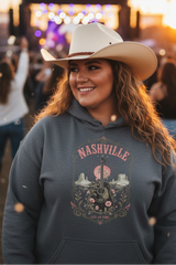 A smiling woman in a cowboy hat and a dark gray graphic hoodie stands outdoors at sunset with a concert stage blurred in the background. The hoodie features a Nashville graphic with a guitar, flowers, and the text 'NASHVILLE' and 'ALL OF THIS'.