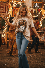 A woman stands outdoors at a festival or party, wearing a beige graphic T-shirt with a gorilla print and the text 'BIG FOOT BELIEVES YOU' paired with blue jeans. The background is filled with warm string lights, tapestries, and other people in a bohemian outdoor setting.