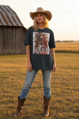 A woman stands in a grassy field wearing a dark charcoal graphic t-shirt with a western rodeo print, light-wash distressed jeans, brown cowboy boots, and a tan cowboy hat. A rustic barn is visible in the background.