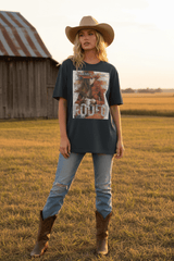 A woman stands in a grassy field wearing a dark charcoal graphic t-shirt with a western rodeo print, light-wash distressed jeans, brown cowboy boots, and a tan cowboy hat. A rustic barn is visible in the background.