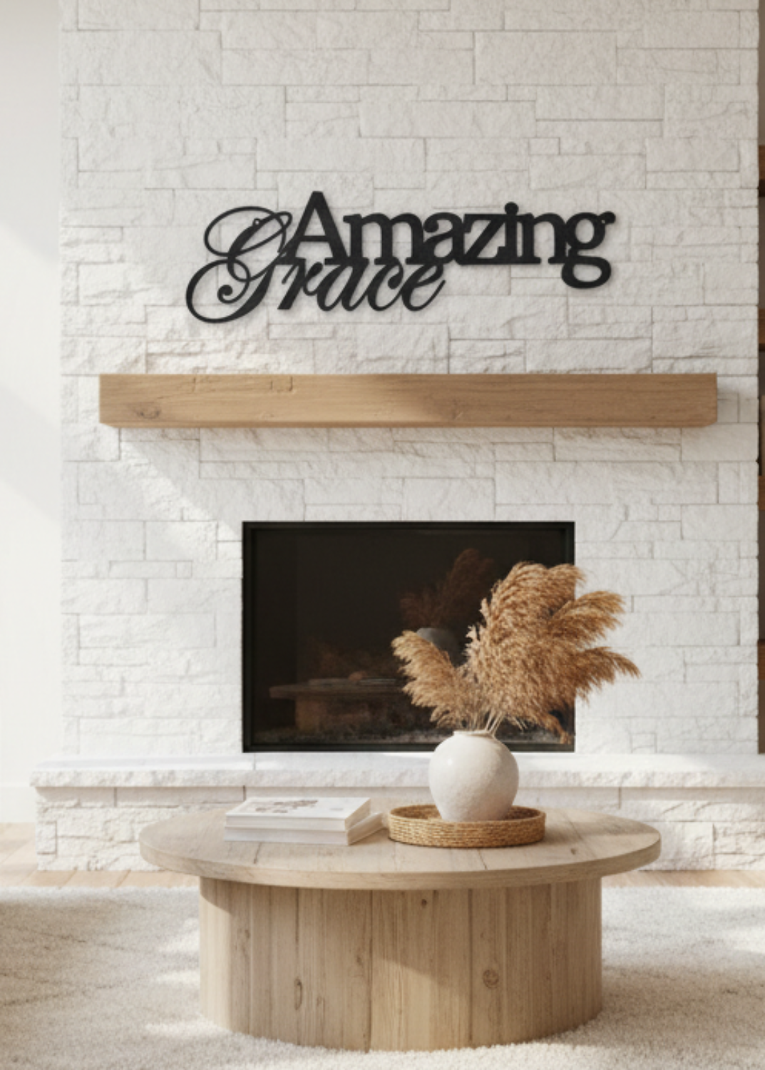 A modern living room with a white brick fireplace featuring black metal wall art that reads 'Amazing Grace'. The room is styled with two light beige sofas, a round wooden coffee table, and built-in wooden shelves on the right.
