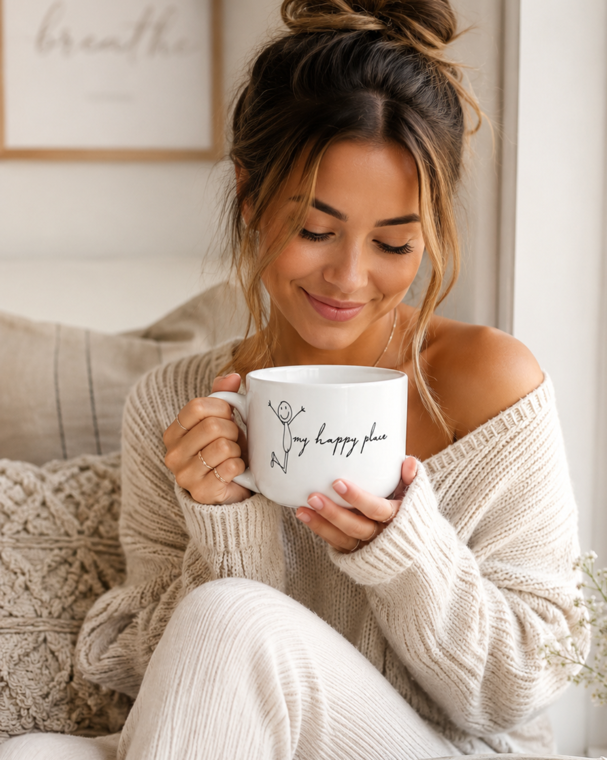 A woman in a cozy off-the-shoulder beige knit sweater and matching pants sits on a couch, holding a white ceramic mug with the words 'My happy place' written in black script. The setting is a bright, warm room with a window, an open book, glasses, and a small vase of baby's breath.