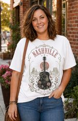 A woman wearing a white graphic t-shirt with a vintage-style 'Nashville' guitar design, paired with blue jeans and a brown shoulder bag, standing outdoors in front of a brick building.