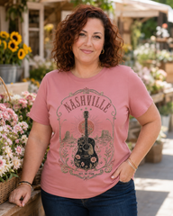 A woman with curly brown hair wearing a dusty pink graphic t-shirt with a black and gray 'Nashville' guitar design, standing in a sunlit garden center with flowers in the background.