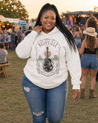 A woman wearing a white Nashville-themed graphic hoodie and distressed blue jeans stands outdoors at a festival, smiling and posing with a crowd and food trucks blurred in the background.