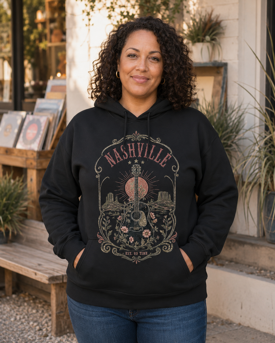 A woman with curly dark hair stands outdoors wearing a black pullover hoodie with a large Nashville graphic on the front, paired with blue jeans. The hoodie features a detailed vintage-style design with a guitar, sun, and floral elements.