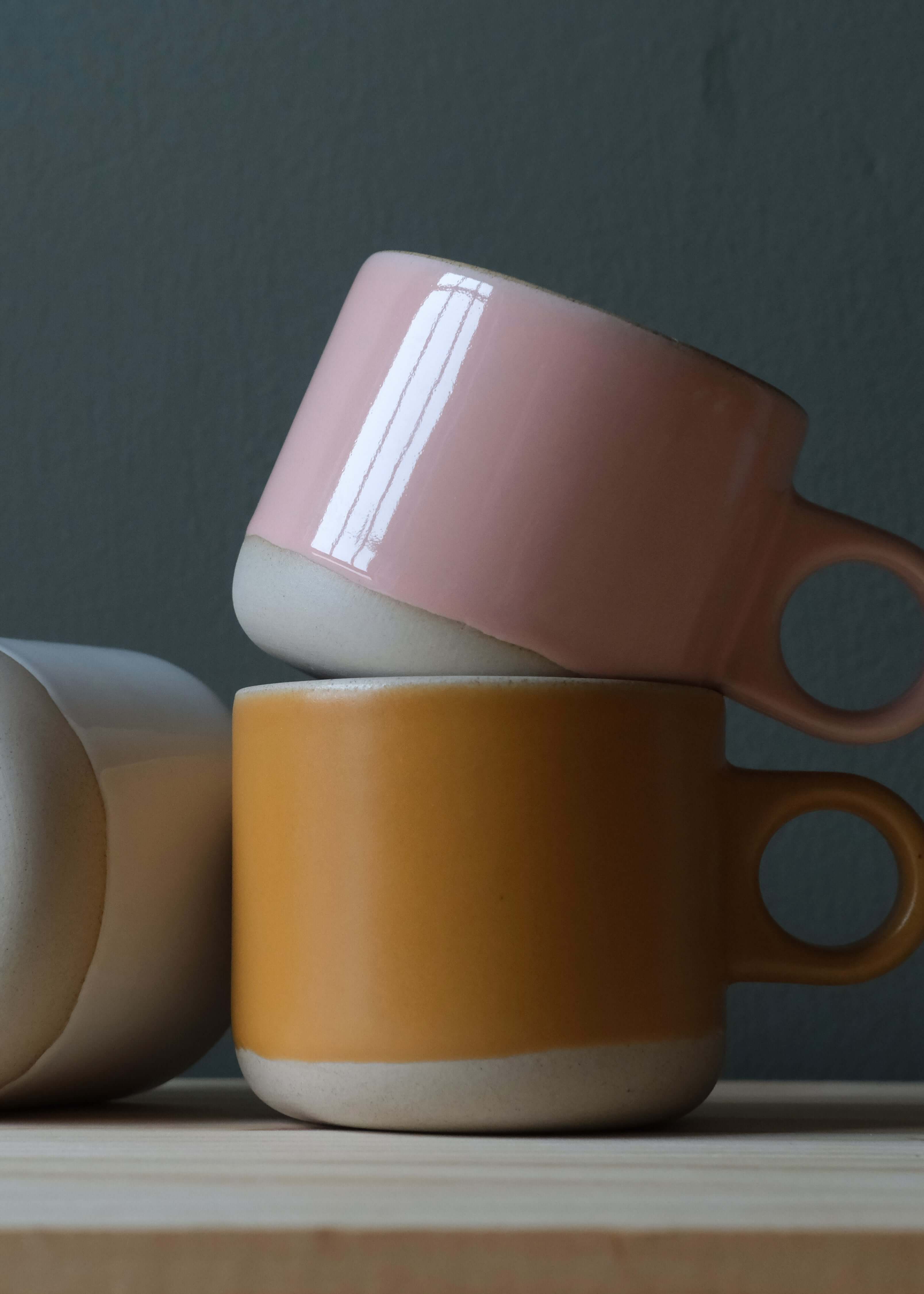 Three ceramic mugs in pink, orange, and beige on a wooden surface with a dark background.