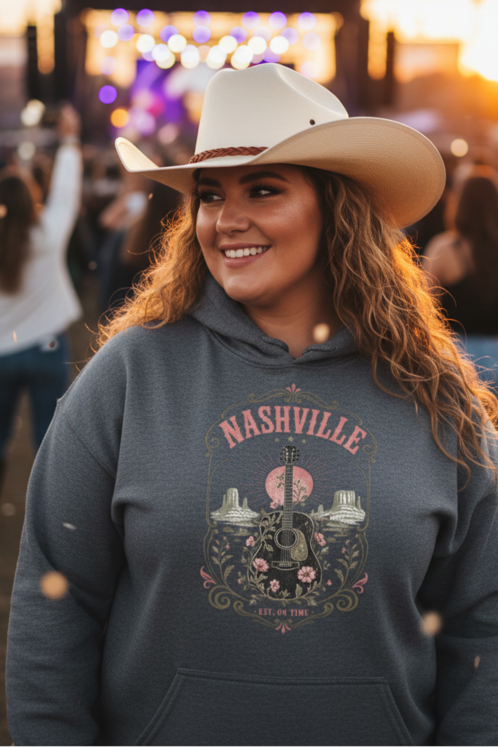 A smiling woman in a cowboy hat and a dark gray graphic hoodie stands outdoors at sunset with a concert stage blurred in the background. The hoodie features a Nashville graphic with a guitar, flowers, and the text 'NASHVILLE' and 'ALL OF THIS'.
